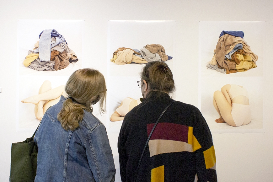 Two people look at six photos of clothing piles and balled up bodies on a white gallery wall