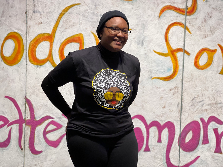 A Black woman wearing all black smiles in front of a painted plywood mural