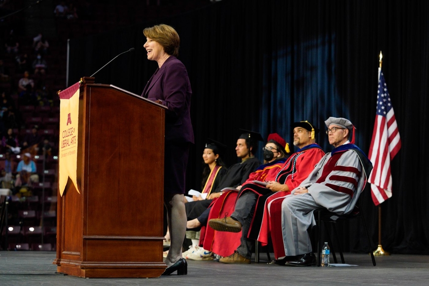 Senator Amy Klobuchar at the podium with several VIPs seated behind her on the commencement stage