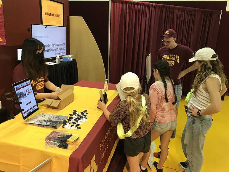 Two statistics students interact with three female fair attendees at a gold and maroon information booth 