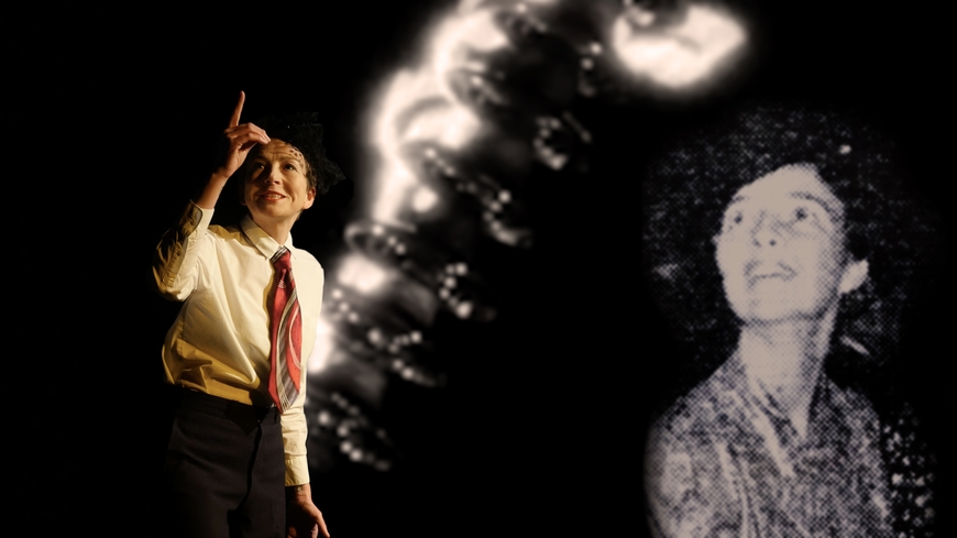 Light-skinned, female-presenting person dressed in a men&#39;s dress shirt and necktie raises finger and looks at it with a smile in from of projected background showing electrical corona discharge and a black and white photograph of another light-skinned, female presenting figure from the 1930s.