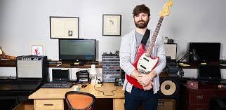 Joel Korte holding an electric guitar standing near a desk with a computer on it