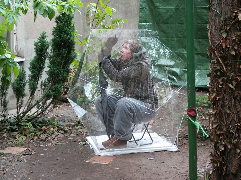 Person sits inside an acrylic polyhedron in a garden, drawing on the inside with a pen