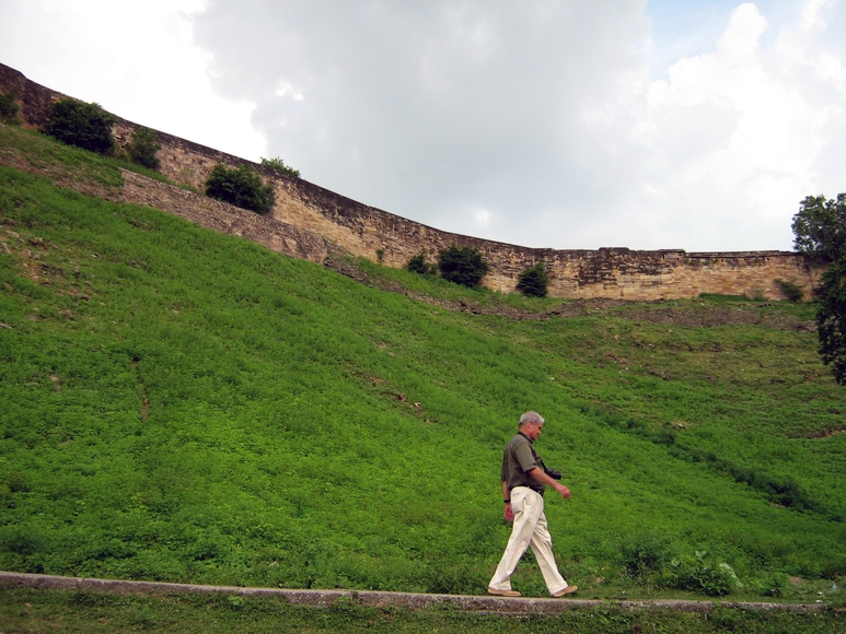 A man walks in front of a steep hill with green grass and a historic fort at the top in India
