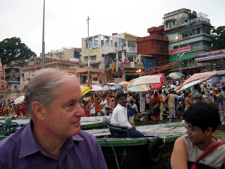 A man and a woman sit in the foreground in a city in India with buildings and crowds behind them