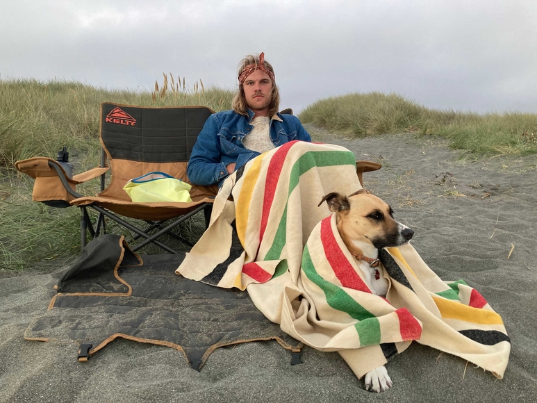 Middle aged man sitting in a camping chair on the beach with a dog lying at his feet.