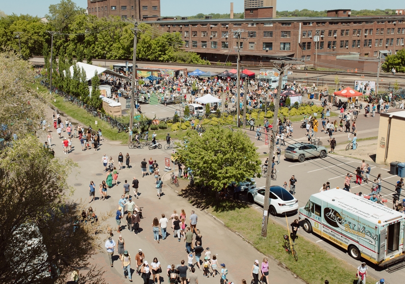 A crowd of people walking past old buildings, food trucks, and vendor tents at an outdoor fair