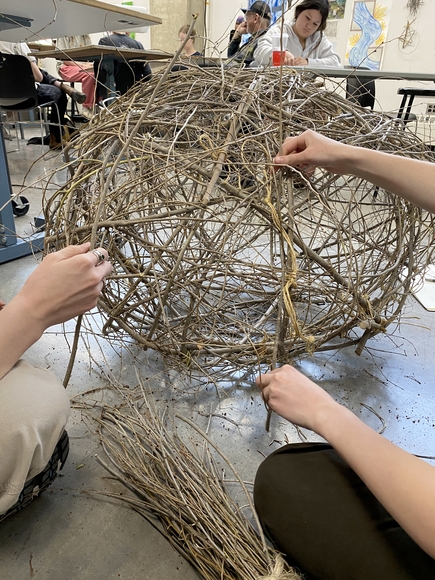 Two people sit on the floor working on a woven orb of sticks. Students work at tables in the background