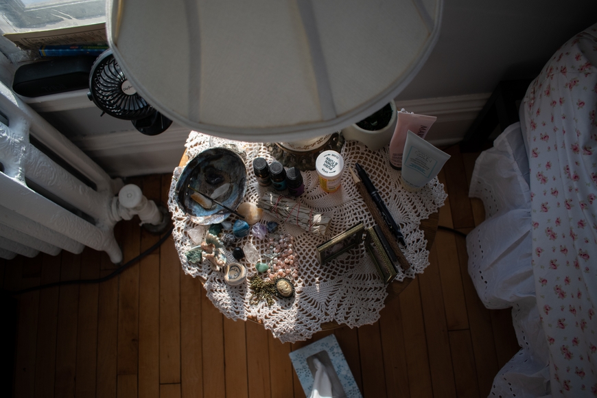 Aerial view of a softly light bedroom side table covered with a lace doily, bottles of oils, medication, and lotions, intermixed with jewelry and other personal items.