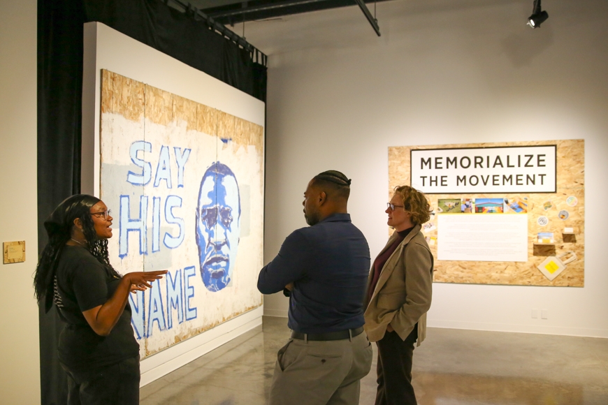 a woman explains a mural in a gallery to two faculty members