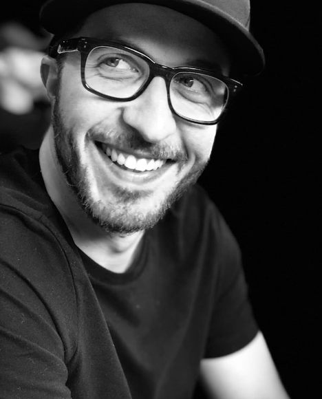 A black and white headshot of Ben Hering, smiling and wearing dark frame glasses
