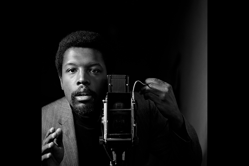 A Black man looks directly at an old fashioned 1960's camera snapping a self-portrait.
