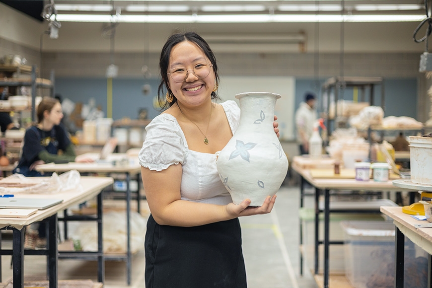 Student smiling at the camera while holding their painted ceramic vase 