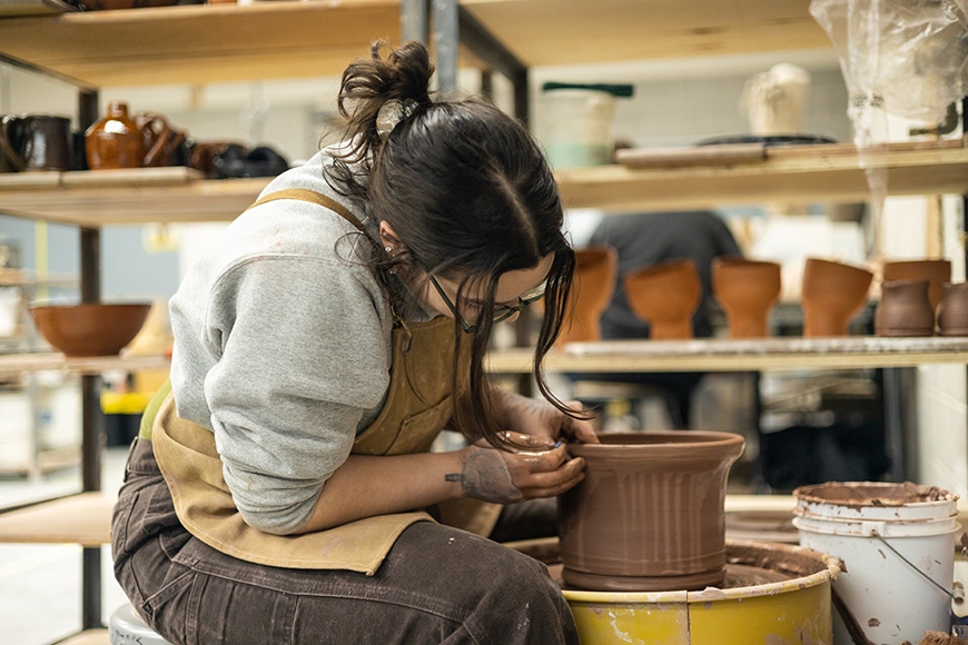 Student in a tan apron sits at the pottery wheel, focused on their project