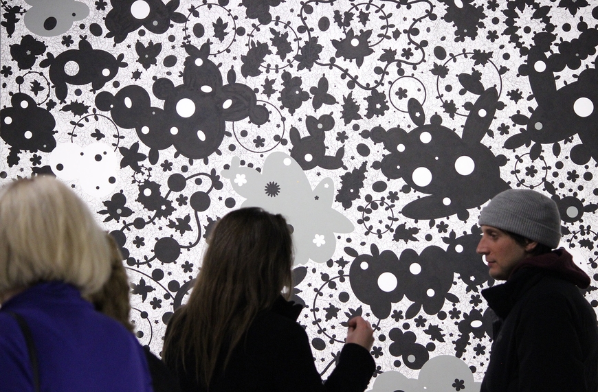 Three people stand talking in front of a giant black and white painting filled with organic circular shapes and marks