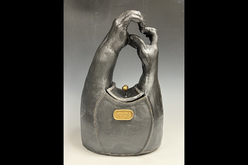 A graphite colored ceramic vessel composed of sculpted human forearms and hands creates a handbag complete with a golden handled inset lid and gold insignia on the front.