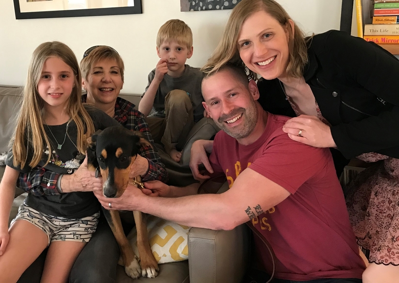 Carrie Strief embraces her husband, alongside two children, another family member, and a dog, who sit on a couch together in a home.
