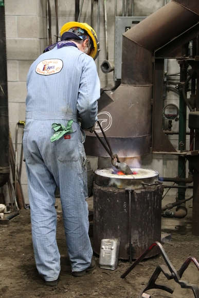 A man in blue jumpsuit and yellow hardhat feeds metal into the furnace with tongs