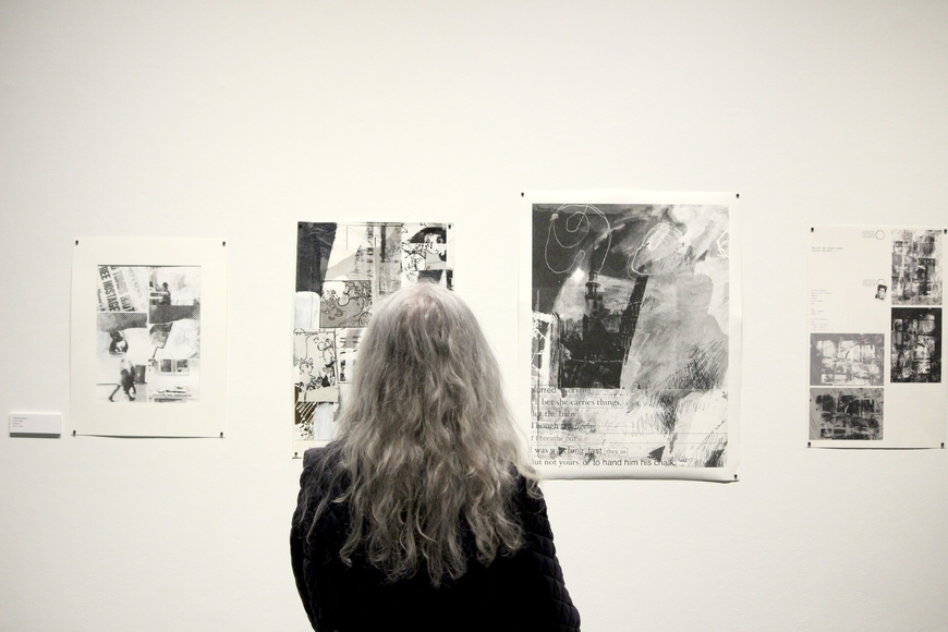 A person with long gray hair looks at black and white mixed media collages on a white gallery wall