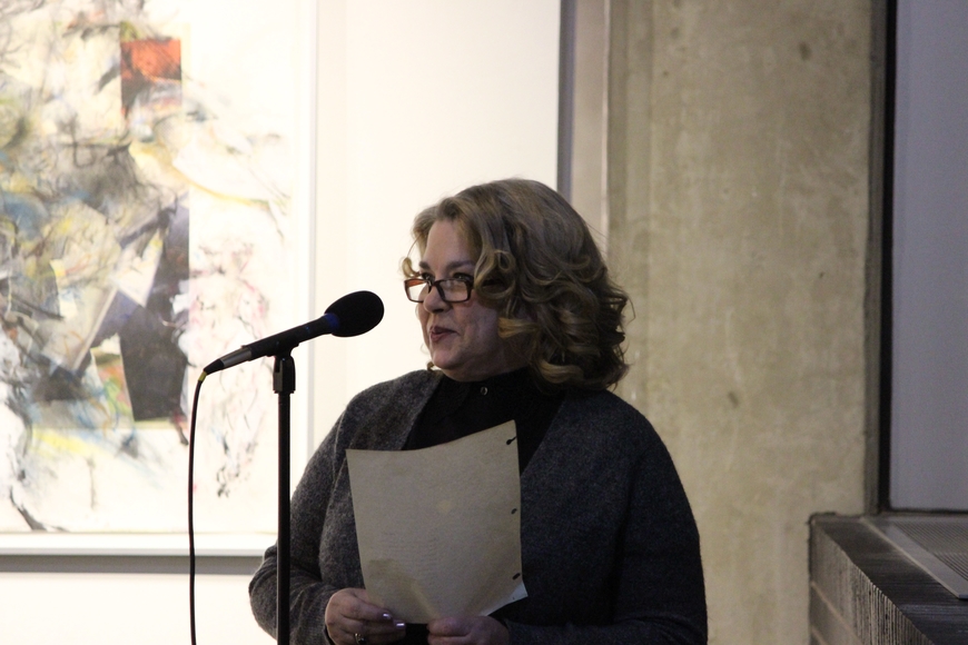 A woman with reading glasses speaks into a microphone in front of abstract art