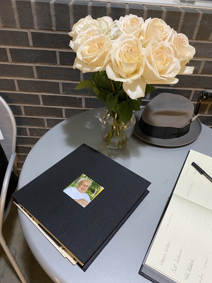 A black book with a man's photo on it sits on a table next to a vase of white roses and a gray fedora