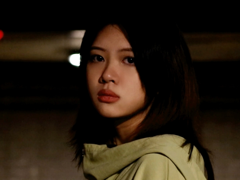 A young person stares directly into the camera with a nightscape in the background.