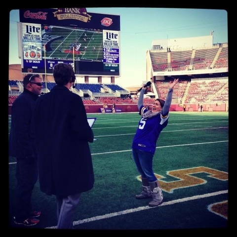 A woman wearing a purple Vikings jersey lifts up her arms triumphantly on a football field