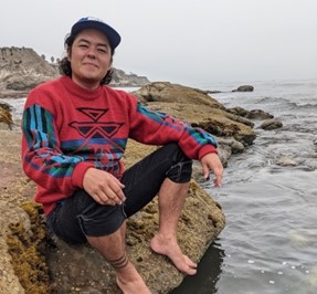 Isaac Espósto in red tribal printed shirt and hat on sitting on a rock in front of a body of water