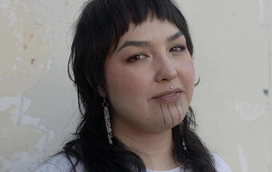 Young Indigenous person with traditional chin tattoos standing against a cream wall