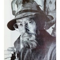 Bearded man in a hat and glasses holds a cigarette