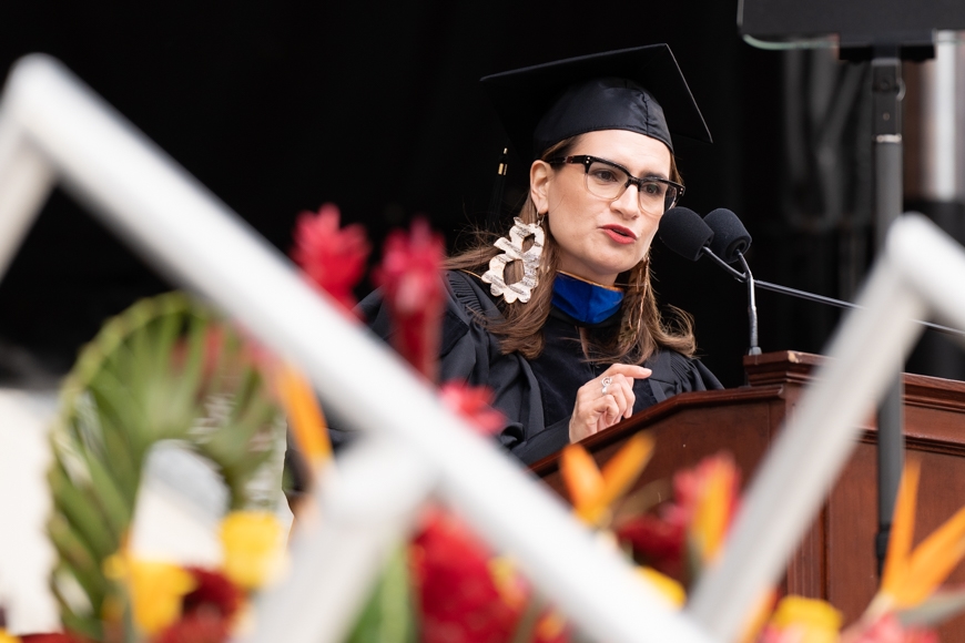 Woman wearing a black cap and gown, glasses, and large earrings speaks at a podium