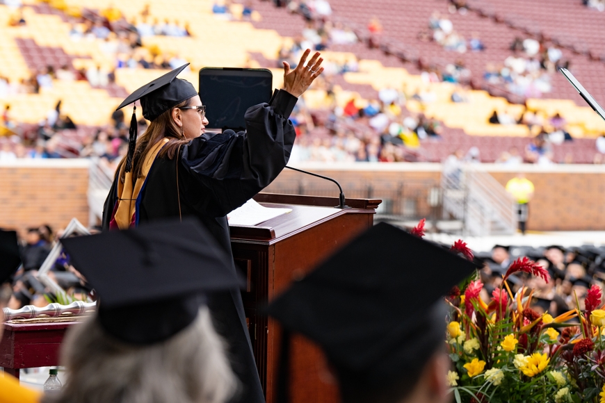 Woman wearing a black cap and gown, glasses, and large earrings is seen from the side as she speaks at a podium
