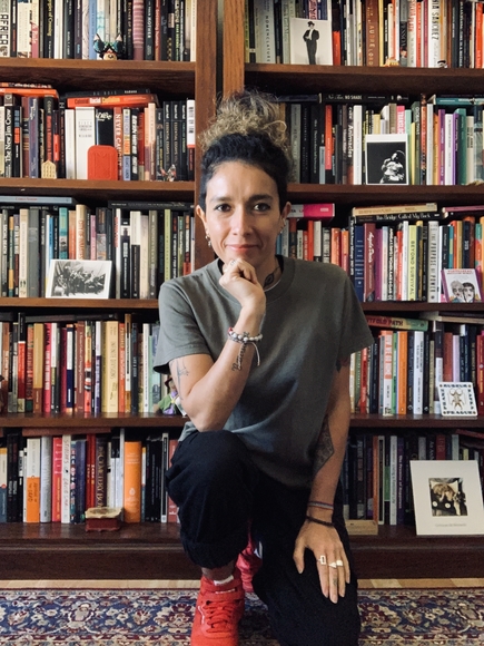 Portait of a person wearing a grey t-shirt, black pants, and red sneakers, with their hair in a bun on the top of their head, kneeling in front of a bookshelf with their head resting in their right hand.  