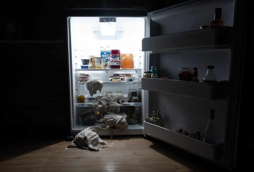 An open refrigerator in a dark room reveals blobs of clay falling out onto the floor