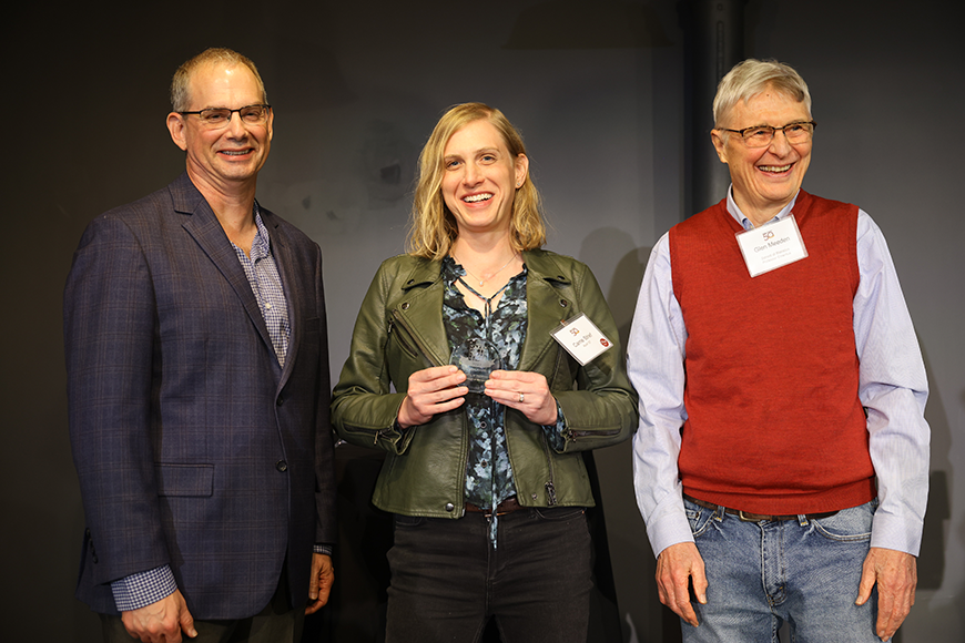 Galin Jones and Glen Meeden stand on either side of Carrie Strief, who is holding a baseball sized, clear glass alumni award. Jones is wearing a suit, Strief is wearing a flowered shirt and green jacket, and Meeden is wearing a collared shirt and sweater vest.