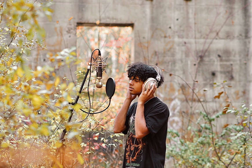 A young man stands in an overgrown landscape while singing into a recording mic as his hand rests on a pair of headphones.