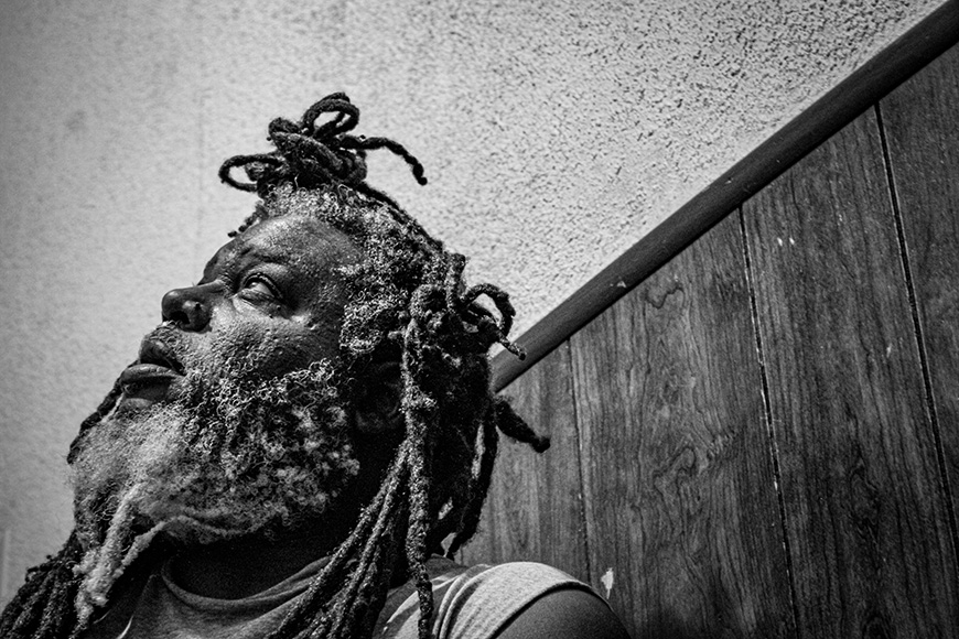 A middle aged man with dreadlocks looks up and off as if transfixed while standing in front of a stucco wall with wood paneled wainscoting.