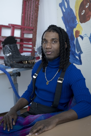 Black man in blue shirt with suspenders sits in an art studio