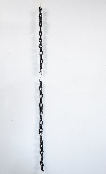 A latex chain coated in asphaltum—a shiny, deep brown black coating—hangs from a white plastic hook against a wall, it has broken a little less than halfway down. The bottom half is adhered to the wall and stays in place.