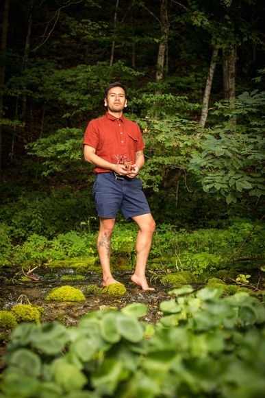 Artist Fidencio Fifield-Perez stands in a forest wearing a red shirt and blue shorts.