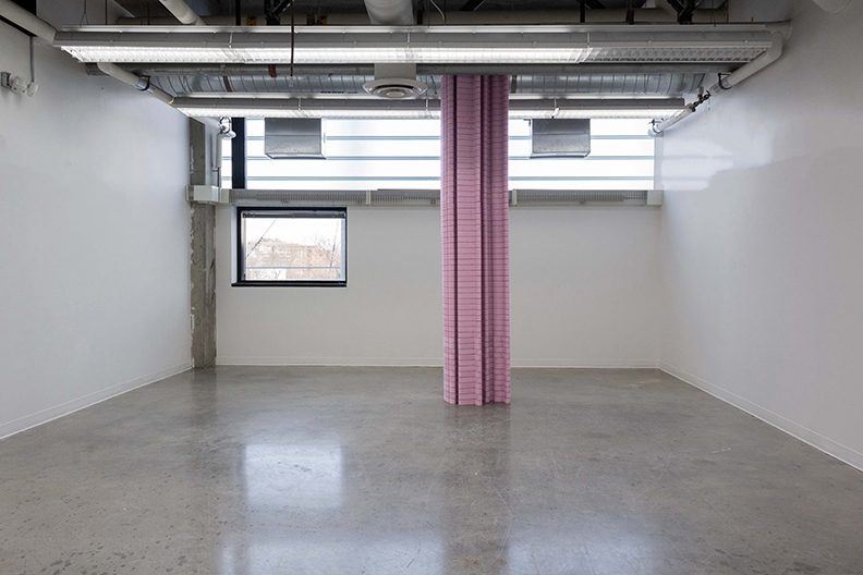 A sculpture made of stacked pieces of pink insulation foam extend up into the industrial ceiling of a room with concrete floors and window on the left hand side.