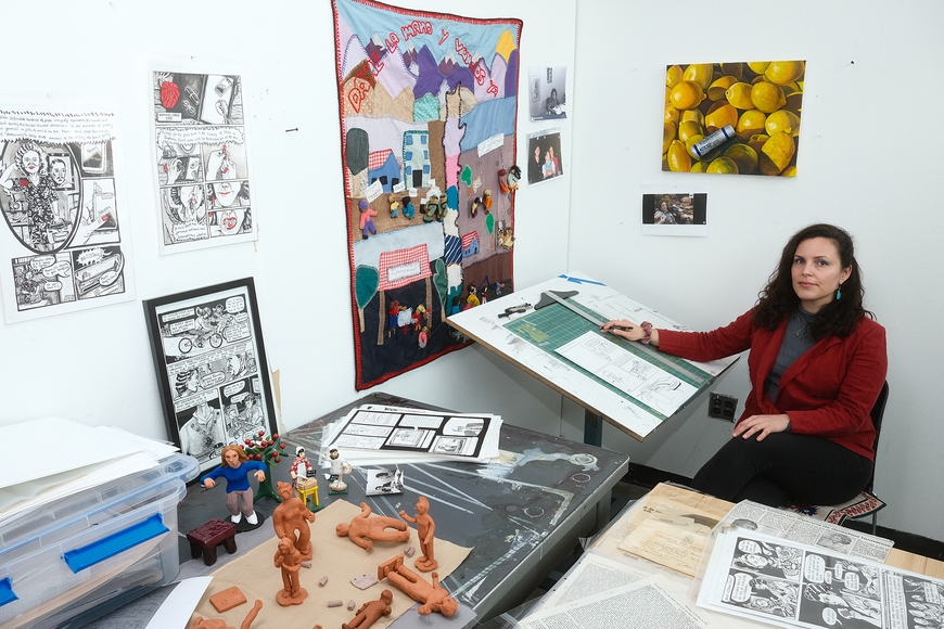 An artist sits at her drawing table in the corner of an art studio with small artworks and figurines on the walls and tables