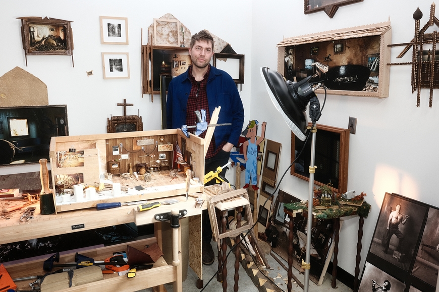 A man stands in an art studio surrounded by wooden dioramas and assemblages