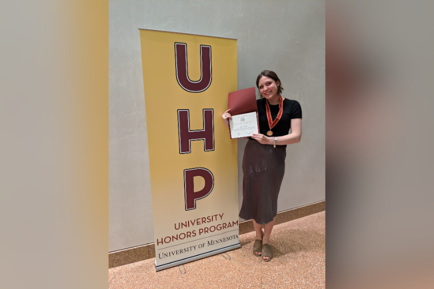 Sophia poses for a picture next to a UHP banner. She is holding her degree