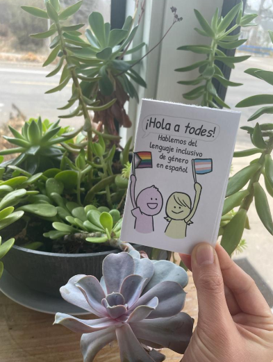 Hand holding a small zine titled ¡Hola a todos! in front of potted plants
