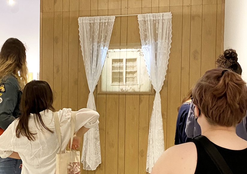 Installation view of wood panel wall with lace curtains and a projection of a closed window. People stand in front looking at the work