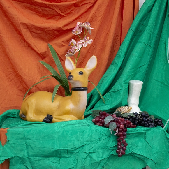 Still life display of plastic deer, grapes, and flowers on orange and green fabrics