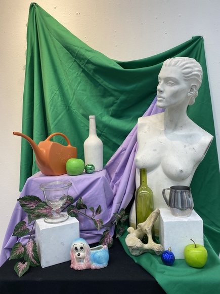 Still life display of mannequin, fruit, vessels, and dog ceramic on green and purple fabrics