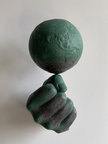 A large right fist balances a small model of the earth on the knuckle of its thumb. Both are textured and colored in green and dark grey to resemble patinated bronze.