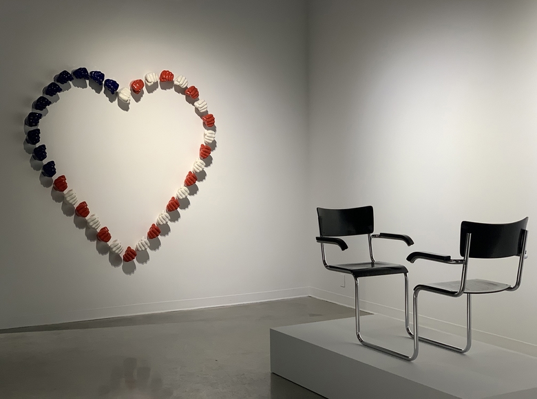 34 ceramic fists painted red, white, and blue attached to the white gallery wall in the shape of a heart, facing in the foreground, on a pedestal, two black wooden office chairs connected by the same bent chrome and facing each other.